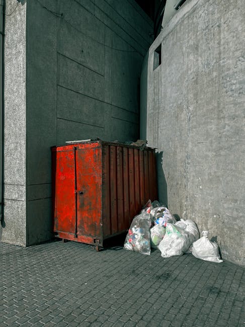 A red metal waste skip with visible signs of rust and chipping paint is positioned against a dark green wall and a light grey concrete wall in an outdoor urban setting. Several plastic garbage bags filled with mixed rubbish are piled at the base of the skip, some leaning against its side. The bags contain various waste items, and the scene is illuminated by natural daylight, highlighting the textured surfaces of the skip, the smooth pavement, and the walls. The pavement consists of dark, interlocking concrete tiles, and the environment appears to be a back alley or side street area, typical for private rubbish collection or waste management services such as those offered by Waste Disposal Colliers Wood, which handles rubbish removal and alternative waste handling options outside standard council collection zones.
