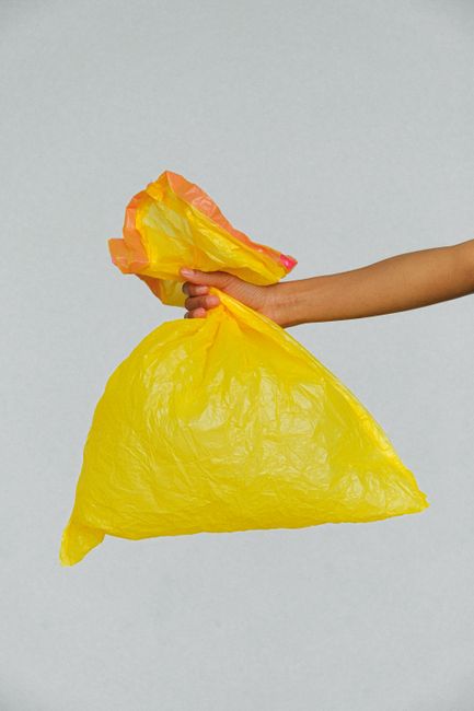 A human hand with a light skin tone is holding a large, bright yellow plastic rubbish bag against a plain light grey background. The bag appears to be filled with waste, with some material visible at the top, including a crumpled orange and yellow plastic sheet. The hand is gripping the bag's opening, and the arm extends from the right side of the image. The scene emphasizes waste disposal and the handling of refuse, aligning with private rubbish collection services offered by Waste Disposal Colliers Wood, such as on-site clearance or alternative waste handling solutions.