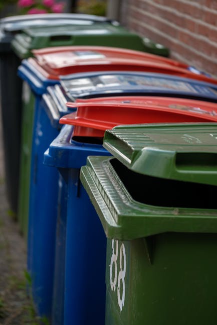 A row of five large, plastic wheelie bins lined up on a paved surface beside a brick wall, with each bin having a different colour lid including green, red, blue, and dark green, all with textured, ridged lids. The bins are positioned next to each other, with the green bin in the foreground on the right and the others receding into the background. The surface is a concrete or tarmac pavement, with some grass or dirt visible at the edges. The scene suggests an outdoor communal or business waste disposal area, where waste collection or private rubbish removal services, such as those provided by Waste Disposal Colliers Wood, might operate, supporting alternative waste management arrangements for shops or local businesses.