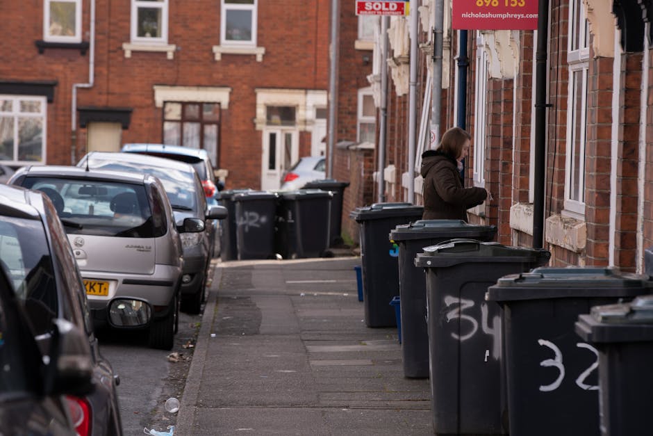A residential street scene showing a row of dark grey plastic wheelie bins placed along the pavement adjacent to red brick terraced houses. The bins, some with lids open or partially closed, are positioned in front of the houses, which have white-framed windows and brickwork detailing around the sills and doorways. A woman dressed in a dark coat is seen at the second house from the right, placing or retrieving items from one of the bins. Several parked cars, including a silver hatchback and a darker vehicle, line the street on the left side, with the pavement extending into the background where additional bins are visible near other houses. The street is outdoors, illuminated by natural daylight, with a modest amount of litter near the curb. This scene reflects aspects of private waste collection practices rather than local authority rubbish collection, aligning with services offered by Waste Disposal Colliers Wood.