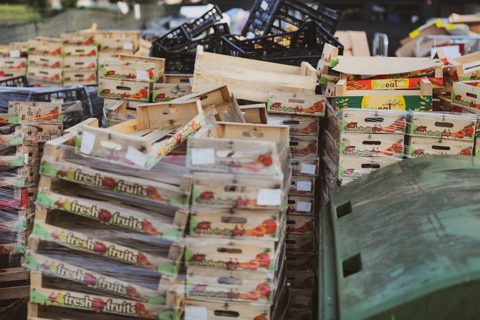 A large collection of wooden fruit boxes stacked haphazardly in an outdoor area, with some black plastic crates mixed in. The boxes are made of light-colored wood with a textured surface, showing visible grain and some minor wear. Each box is decorated with colorful printed labels featuring images of fresh fruits and the words 'fresh fruits' on the sides. The boxes vary in orientation, with some placed upright and others lying on their sides or stacked unevenly, creating a cluttered scene. In the foreground, a rusty, green metal object, possibly a lid or part of a container, partially covers the bottom right corner of the image. The background is slightly blurred, but additional crates and outdoor environment elements are visible, indicating an open storage or waste collection site. The lighting is natural and diffuse, emphasizing the textures of the wood and materials involved, consistent with a scene where waste or surplus materials are gathered for disposal or recycling, reflecting services offered by Waste Disposal Colliers Wood managing waste via independent collection or external disposal methods.