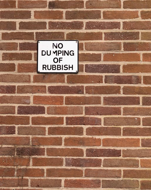 A close-up image of a brick wall featuring a rectangular white sign with black text that reads 'NO DUMPING OF RUBBISH'. The brick wall consists of horizontally laid reddish-brown bricks with a textured surface and light-colored mortar joints. The sign is mounted near the upper left section of the wall, slightly offset from the center, and is framed with a thin black border. The lighting appears natural, casting a subtle shadow to the lower left of the sign, emphasizing the texture of the bricks. This scene depicts a typical exterior setting where waste management or rubbish removal services might be relevant, illustrating a common form of private or non-local authority waste prevention and disposal signage often seen in areas requiring clear communication about rubbish collection policies. Waste Disposal Colliers Wood provides professional rubbish removal services that align with enforcing such local rules and promoting proper waste handling.